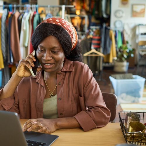Black woman young adult sitting at desk talking on smartphone while using laptop in thrift shop, jewelry and accessories displayed on table, clothing racks visible in background